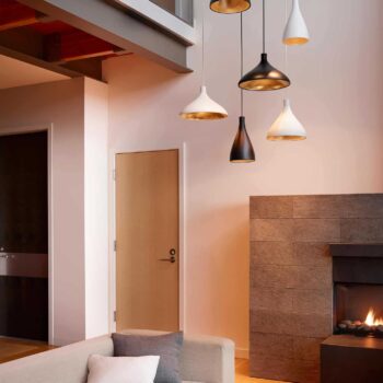 A sophisticated modern living room featuring the Swell Single sectional sofa in beige, accentuated by multi-tone pendant lights and a lit fireplace. The space is enhanced by high ceilings and contemporary decor.