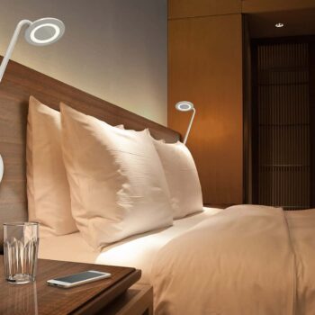 A neatly made bed with two modern wall lamps above the headboard. A wooden nightstand, similar to the Pixo Plus Table, holds a smartphone and a glass of water.