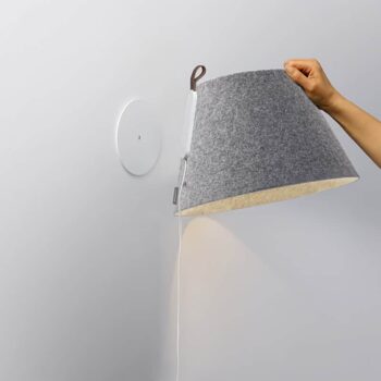 A person adjusts the Lana Floor lamp, featuring a gray felt shade with a white base and power cord, against a light gray wall.