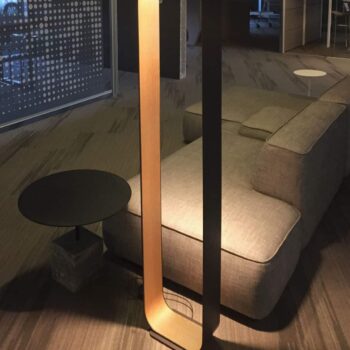 A sleek Contour Floor lamp stands gracefully beside a gray sectional sofa and round side table in the dimly lit room.