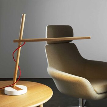 Clamp wooden modern desk lamp with a red cord on a round table next to a gray cushioned armchair against a plain background.