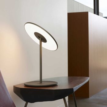 The Circa Table lamp, with its circular flat light, stands on a wooden table. A geometric chair sits nearby as natural light from large windows brightens the space.