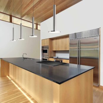 A modern kitchen featuring a large Cielo XL island with a black countertop, stainless steel refrigerator and oven, overhead pendant lights, and light wood floors and cabinets that enhance its minimalist design.