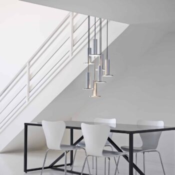 A minimalist dining area with a black table, four white chairs, Cielo Pendant lighting, and a staircase with a white railing as the backdrop.