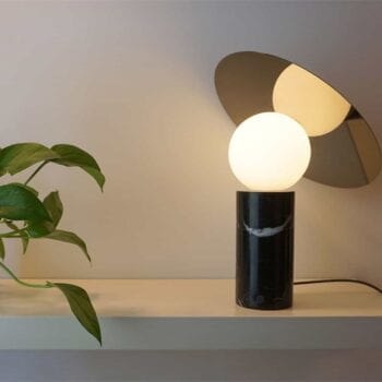 The Bola Disc Table lamp, featuring a modern round bulb and a black cylindrical base, is placed on a shelf beside a small potted plant with green leaves.