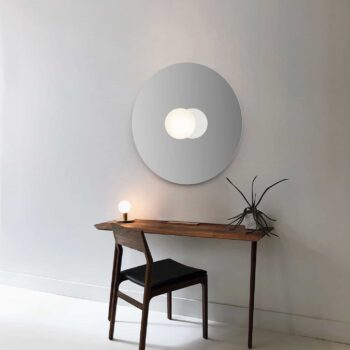 Minimalist room featuring a wooden desk with a chair, adorned by a round mirror and the Bola Flush lamp boasting a small glowing bulb. A plant in a glass container sits beside the lamp.