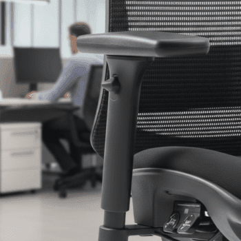Close-up of the C19 Chair’s adjustable armrest, with a person working at a desk in the blurred background of a modern office.