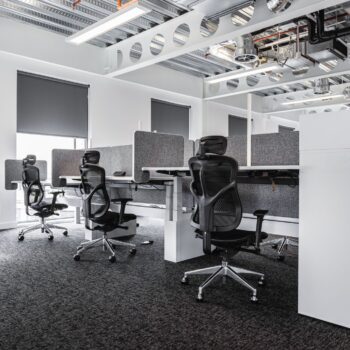 Modern office space featuring several empty workstations, gray partitions, exposed ceiling pipes, and ergonomic F94 Chairs.