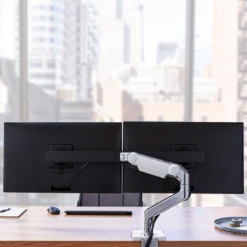 A modern office desk features dual monitors mounted on the M8.1 Monitor Arm Configurator