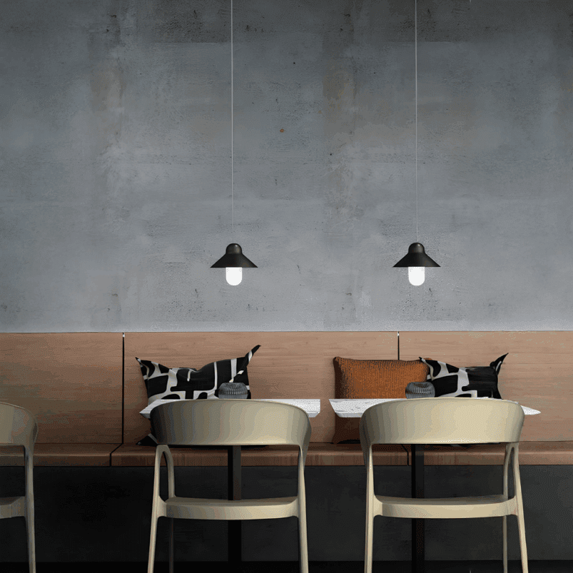 Minimalist café seating area with a light wooden bench, two tables, modern chairs, patterned cushions, and pendant lights creating a warm ambience against a gray wall.