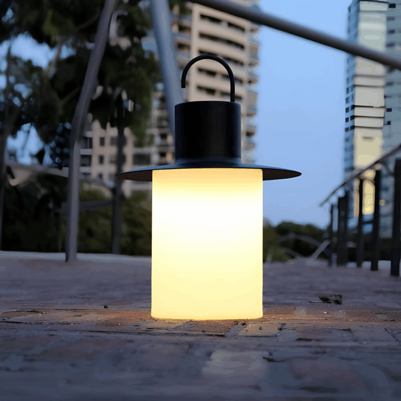 A modern lantern with a black handle and hat-shaped top emits a warm light on a brick pathway, creating cosy ambience for autumn evenings amid tall buildings and trees in the background at dusk.