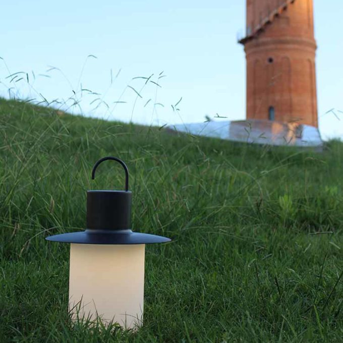 A black lantern with a frosted glass body sits on green grass, adding ambience and warmth to the scene, with a brick tower and blue sky visible in the background.