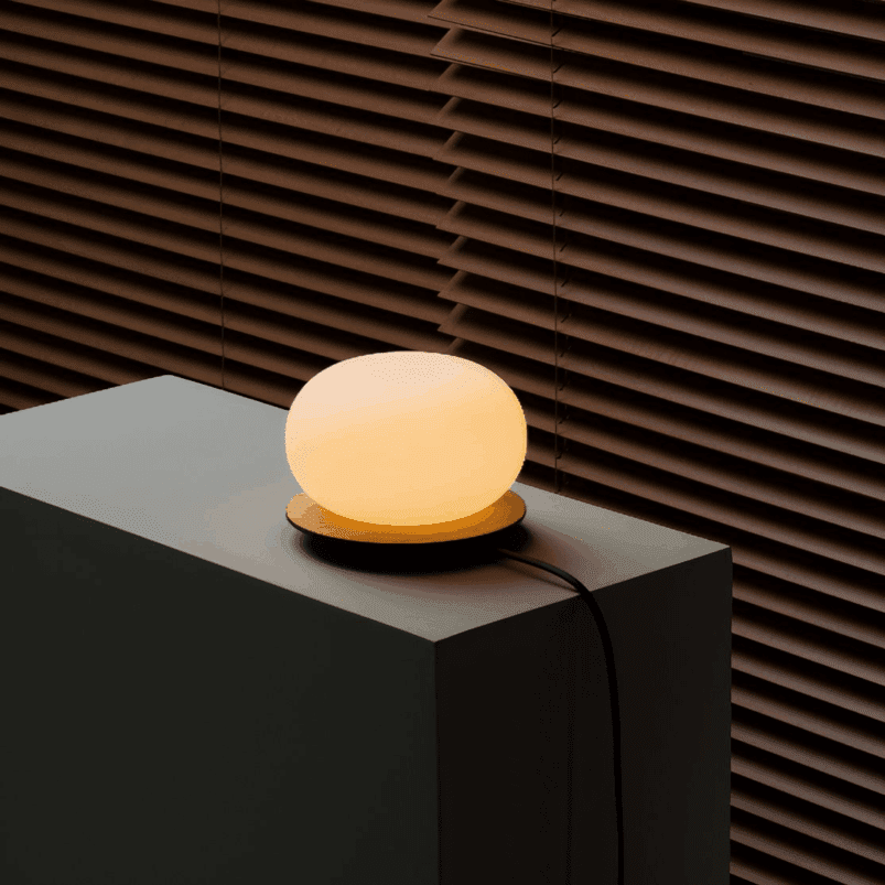A round, glowing lamp sits on a gray pedestal next to a window with closed brown blinds, casting a warm ambience throughout the room.