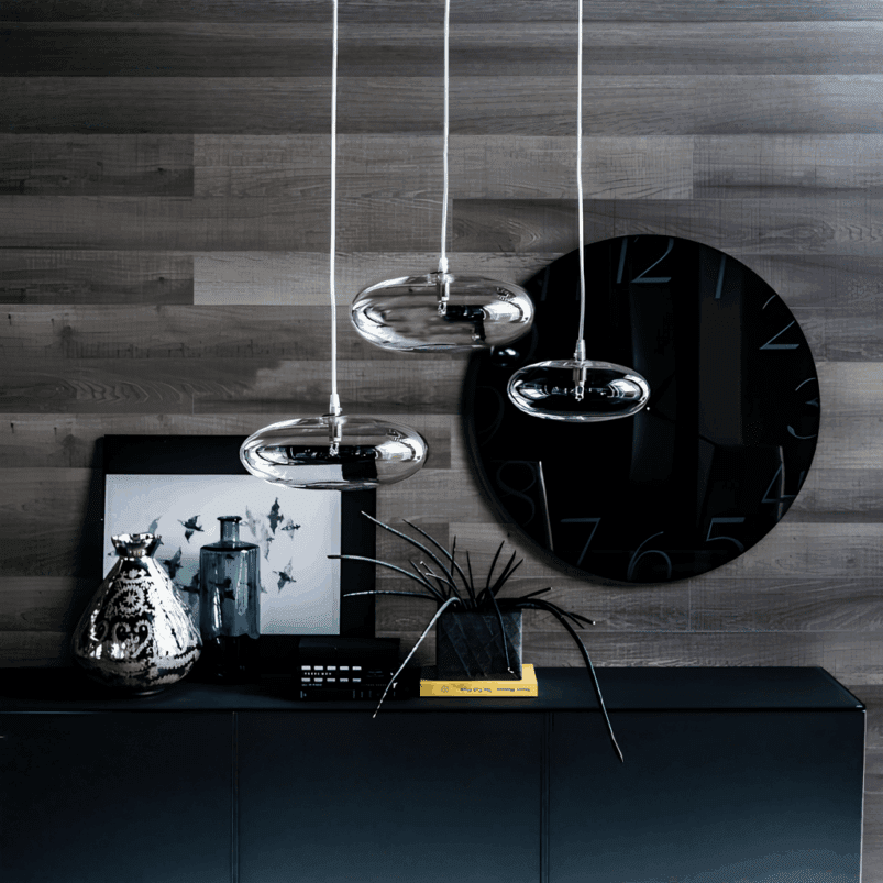 Modern interior with a large black wall clock, decorative vases, a framed photo, and a plant. Three pendant lights create warm ambience and perfect lighting for autumn on a dark cabinet against a grey wood-paneled wall.