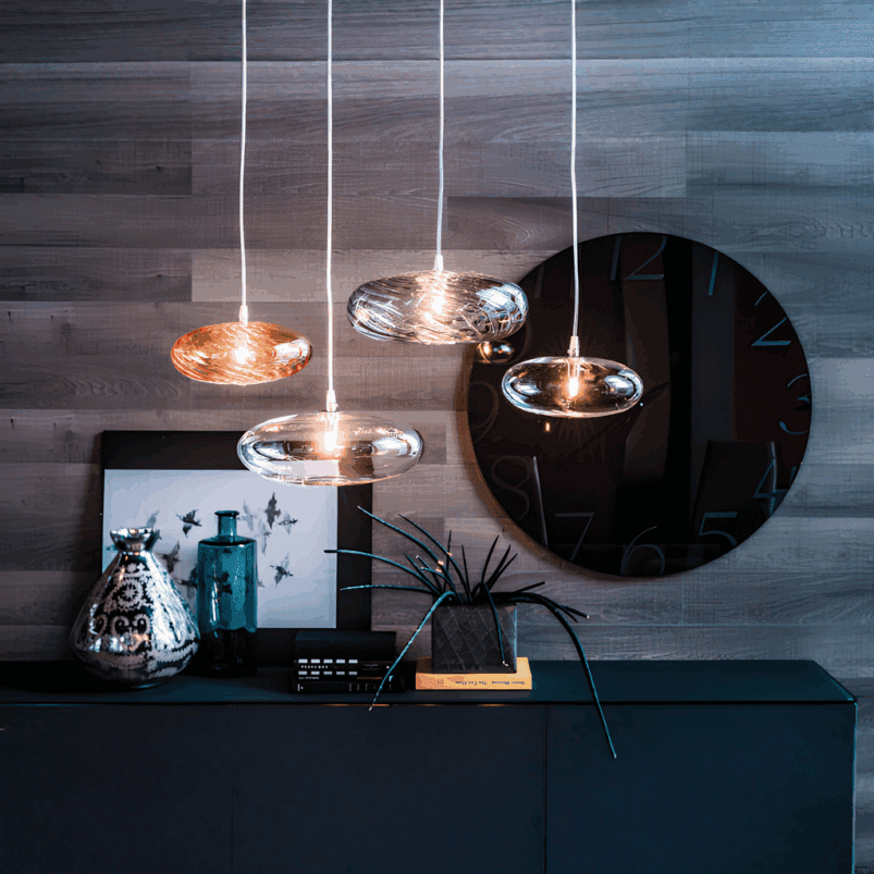 Four modern pendant lights create autumn lighting above a cabinet adorned with a reflective vase, blue bottle, framed art, and a large round black wall clock, adding warmth and ambience to the space.