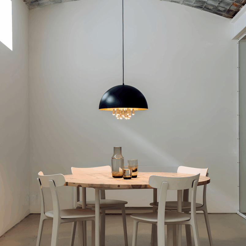 A round wooden dining table with four white chairs, glassware on top, and a black pendant light hanging above creates a minimal, modern room filled with warmth and inviting ambience.
