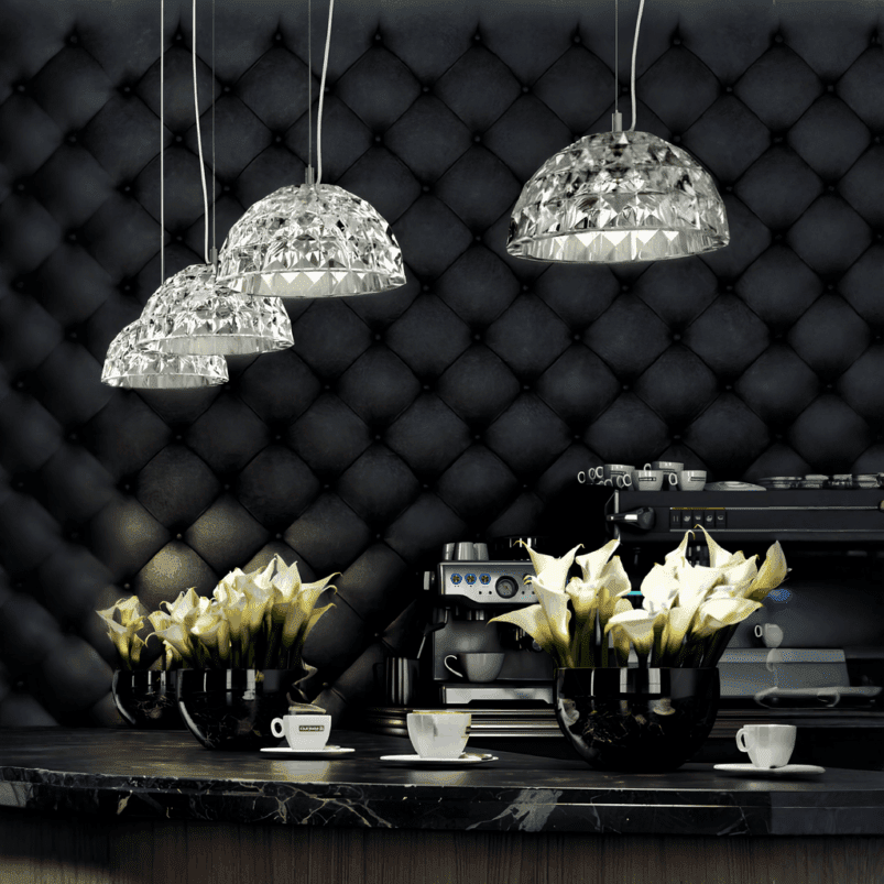 Three pendant lights cast autumn lighting above a counter with coffee cups, a coffee machine, and vases of white calla lilies, set against a dark, tufted wall to create an inviting ambience.