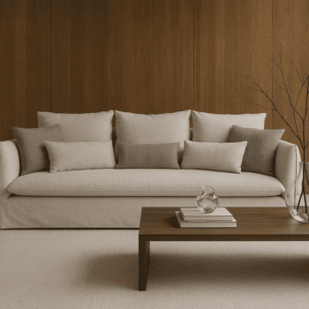 A modern living room featuring the Dylan Sofa in beige with six cushions, a wooden coffee table displaying books, a glass sculpture, and a vase of bare branches, all set before a wood-paneled wall.