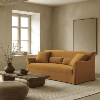 A minimalist living room featuring the Ralph Sofa in mustard, a wooden coffee table, round ottoman, neutral rug, and a large window filling the space with natural light.
