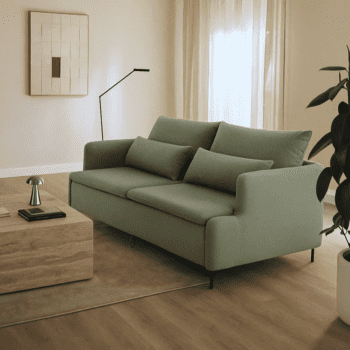 A modern living room featuring the Tower Sofa-bed in light green, a wooden coffee table, black floor lamp, potted plant, and abstract wall art on a light wood floor with neutral decor.