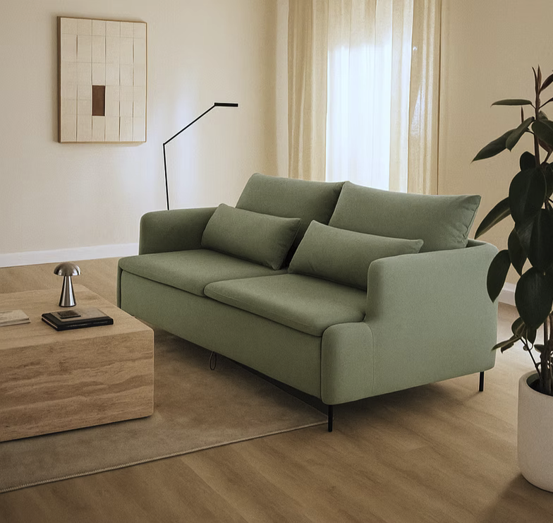 A modern living room featuring the Tower Sofa-bed in light green, a wooden coffee table, black floor lamp, potted plant, and abstract wall art on a light wood floor with neutral decor.