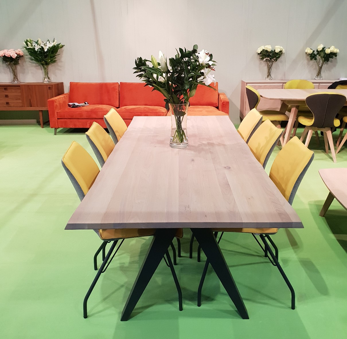 The LOOP Delta table, paired with six yellow chairs and a vase of white flowers on a green floor, stands out with an orange sofa and other stylish furniture in the background—ideal for gatherings or lively LOOP conversations.