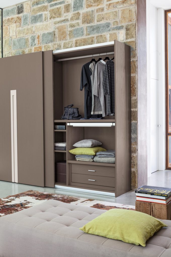 A modern wardrobe with sliding doors is open, displaying hanging clothes, shoes, folded blankets, and drawers inside—a perfect way to furnish a bedroom. A tufted bench sits in front, with a stone wall accenting the finished bedroom décor.