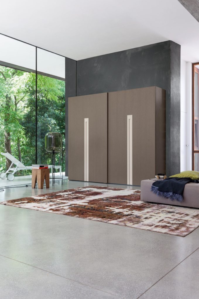 Modern living space ideal for bedroom decorating, featuring large windows, a brown wardrobe, patterned rug, grey sofa, small tables, and greenery visible outside.