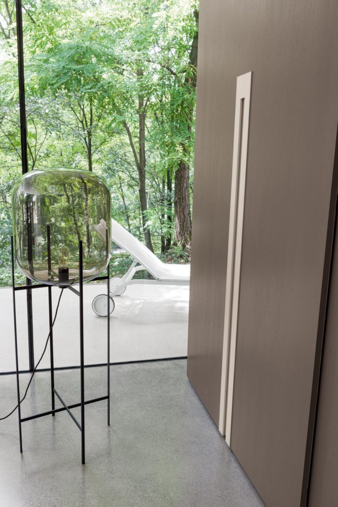 A modern interior with a glass lamp on a black stand, large window revealing green trees, and a white lounge chair outside on a patio—a perfect inspiration for stylish bedroom décor.
