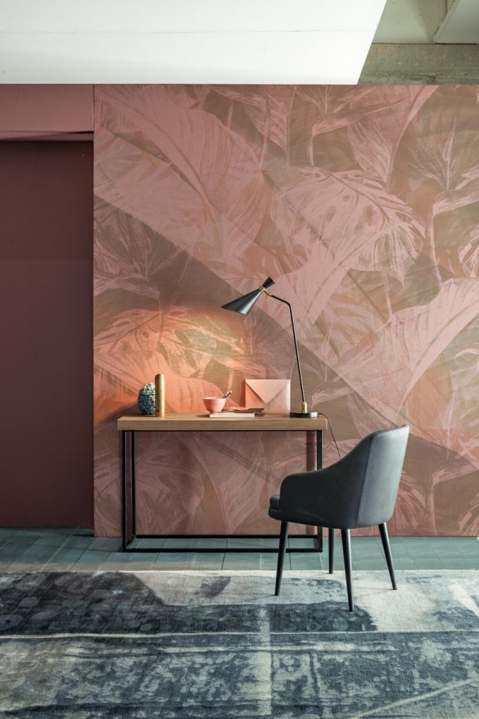 A modern desk with books, a lamp, and décor sits against a patterned pink wall; this chic setup is perfect to furnish a bedroom, while a grey chair and rug complete the minimalist workspace.