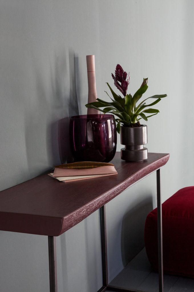 A narrow table with a purple glass vase, a potted plant, and a closed book against a light grey wall adds a modern touch to finished bedroom décor.