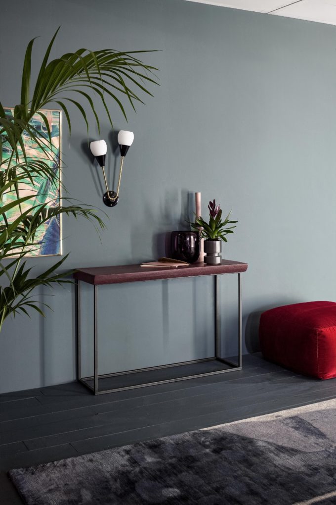 A modern entryway with a slim console table, decorative vases, a wall sconce, a red pouffe, and framed artwork against a muted green wall—perfect inspiration to furnish a bedroom or elevate your finished bedroom decor.