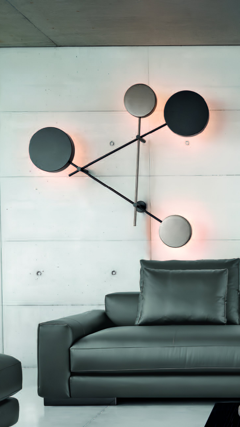 Modern wall-mounted circular light fitting with black and grey discs creates striking bedroom décor above a grey leather sofa with matching cushions in this minimalist finished bedroom.