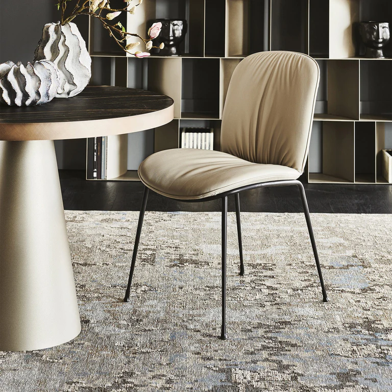 A modern beige chair with black metal legs sits next to a round table on a patterned rug, perfect to furnish a bedroom, with shelves and decorative items in the background for stylish bedroom décor.