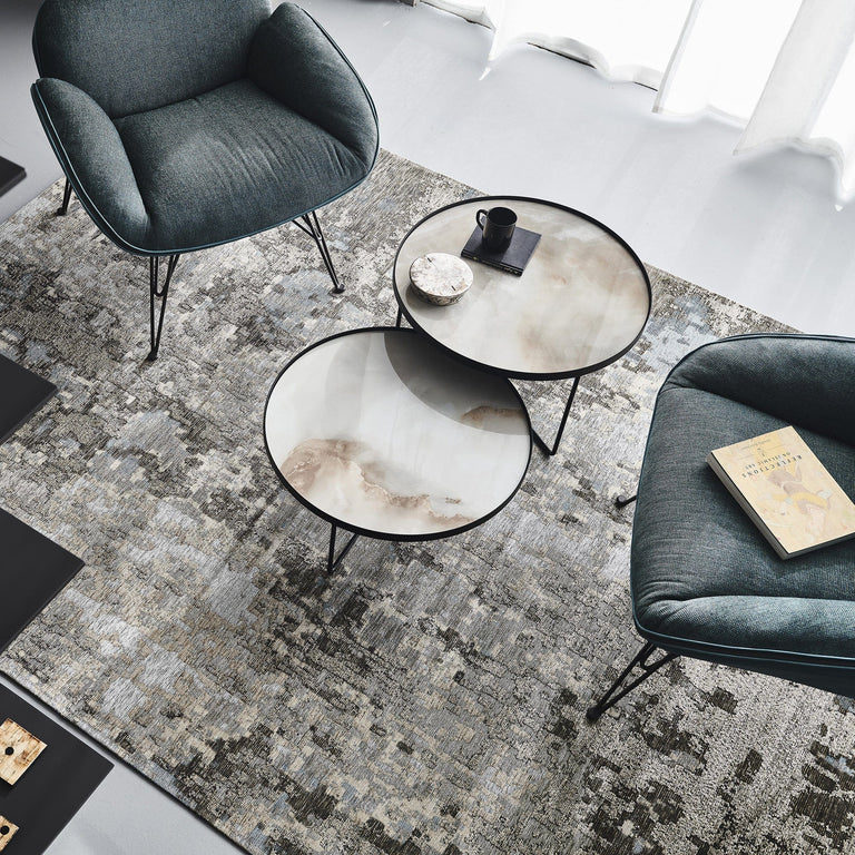 Two modern armchairs and nesting round coffee tables sit on a patterned rug; one table holds a cup and saucer, while the other has a closed book—perfect touches to inspire your finished bedroom or elevate your bedroom décor. Light streams in from nearby windows.