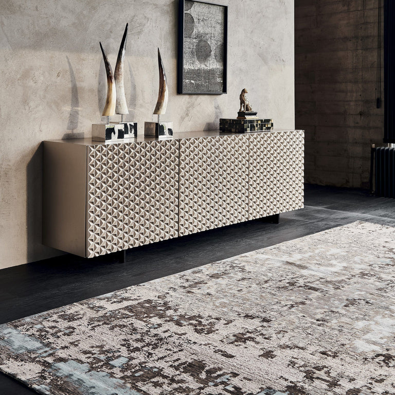 Modern sideboard with a geometric textured front stands against a concrete wall, decorated with abstract sculptures, a framed artwork, and a small figurine on a patterned rug—perfect to furnish a bedroom with contemporary style.